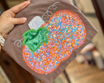 Op de afbeelding: Een bruin T-shirt met een applicatie van een pompoen met pailletten. De pompoen is oranje met iriserende pailletten en een groene fluwelen steel.