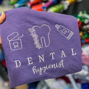 Op de afbeelding: Paars T-shirt met witte borduursels van een tand, een tube tandpasta en een flosdraaddoosje. De tekst "DENTAL hygienist" is onder de afbeeldingen geborduurd.