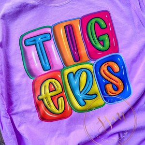 May include: A purple t-shirt with the word "TIGERS" printed in colorful, outlined letters that resemble inflated balloons.