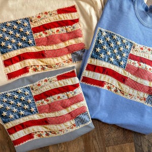May include: Three crewneck sweatshirts in cream, light blue, and grey, each featuring a large, rectangular American flag applique. The flag is constructed from various fabric strips in red, white, and blue, including floral and gingham patterns.