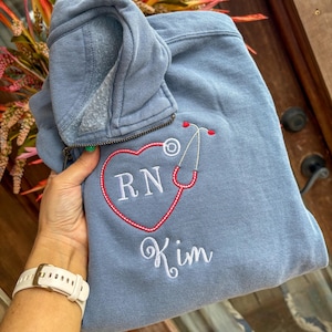 May include: A dusty blue quarter-zip sweatshirt featuring an embroidered design of a red heart shaped like a stethoscope. Inside the heart, the letters RN are stitched in white, and the name Kim is embroidered in white script below the heart.