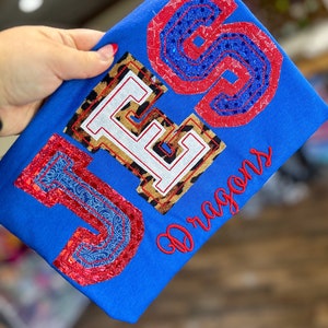 May include: Blue t-shirt with the letters "JES" in different colors and patterns. The letter "J" is blue and red with a paisley pattern, the letter "E" is brown and white with a leopard print, and the letter "S" is red and blue with a star pattern. The word "Dragons" is embroidered in red below the letters.