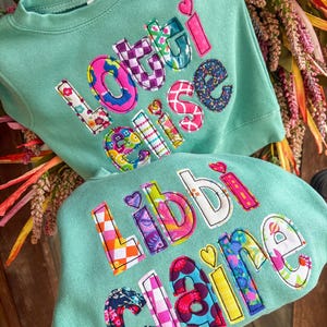 May include: Two mint green sweatshirts featuring colorful, patterned fabric applique letters. The top sweatshirt displays the name Lottie Elise, and the bottom sweatshirt displays the name Libbi Claire. Each letter is outlined with black stitching.