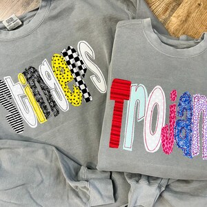 May include: Two gray sweatshirts with colorful patchwork lettering. The first sweatshirt says "Stingers" in yellow, black, white, and gray letters. The second sweatshirt says "Trojans" in red, blue, pink, white, and gray letters.
