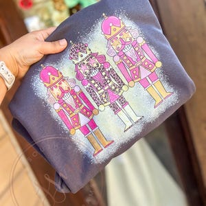 May include: A gray sweatshirt with a design of three pink and gold nutcracker figures. The nutcrackers are wearing pink and gold hats and have gold accents. The middle nutcracker is wearing a leopard print outfit.