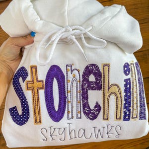 Puede incluir: Una sudadera con capucha blanca que presenta la palabra Stonehill aplicada en letras de tela con patrones morados y dorados, con una cruz integrada en la letra t. Debajo del texto principal, la palabra skyhawks está bordada en fuente negra.