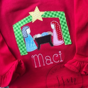 May include: Red long-sleeved shirt with a green, yellow, and pink applique of the nativity scene. The shirt has a ruffled trim around the neckline and sleeves. The name "Maci" is embroidered below the applique.
