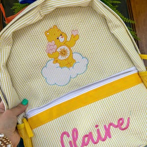 May include: A yellow and white pinstriped backpack featuring an embroidered yellow Care Bear character sitting on a white cloud, holding a pink heart. The front pocket displays the name Claire embroidered in bright pink cursive script lettering.