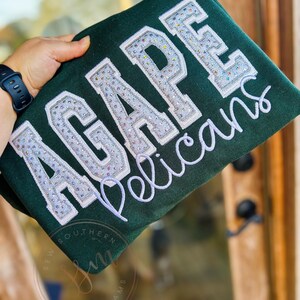 以下が含まれることがあります： 緑色のスウェットシャツに、白と銀のリボンで「AGAPE Pelicans」と書いてあります。スウェットシャツの左下には、「Southern Sew & Monograms」という小さなロゴがあります。