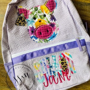 Peut inclure: Un sac à dos rayé violet et blanc avec une applique de cochon fleurie et le nom "S. Jane" brodé sur le devant.