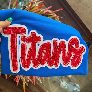 May include: A bright blue sweatshirt featuring the word Titans in large, red, sequined letters. The text is outlined in a thick white border, creating a bold contrast against the blue fabric. The lettering has a textured, shimmering appearance.