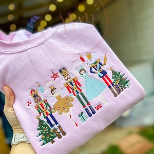 Puede incluir: Sudadera rosa con un diseño bordado de un Cascanueces, una bailarina, un rey ratón y un árbol de Navidad. El diseño es de estilo caprichoso con detalles coloridos.