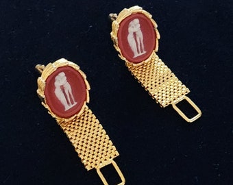 May include: A pair of gold cufflinks with red cameo insets featuring a white silhouette of two figures embracing. The cufflinks have a chain-link design.