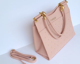 May include: A blush pink handbag with gold-tone hardware and a detachable shoulder strap. The bag features a braided handle and small, evenly spaced studs. The brand name "bebe" is visible on the front.