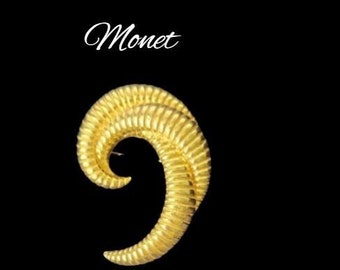 May include: A gold-toned, abstract, swirl-shaped brooch with the word "Monet" written above it.