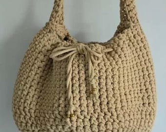 May include: A beige, handmade crochet bag with long shoulder straps. The bag has a drawstring closure with a decorative knot and small gold-colored beads. The bag is hanging on a white hanger against a plain background.