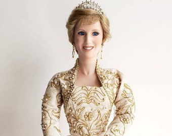 May include: A porcelain doll dressed in a white gown with gold embroidery and a pearl tiara. The doll has blonde hair and blue eyes. The doll is holding a tag that says "Diana, Princess of Wales".