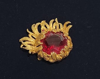 May include: A gold-toned brooch with a red gemstone in the center, surrounded by a decorative design of gold leaves.