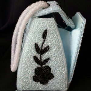 May include: A light blue beaded handbag with a black floral design. The bag has a white beaded handle and a flap closure.