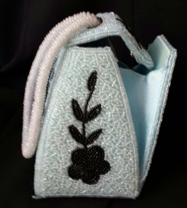 May include: A light blue beaded handbag with a black floral design. The bag has a white beaded handle and a flap closure.