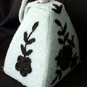 May include: A light blue beaded handbag with a black floral design. The bag has a handle and is shaped like a pyramid.