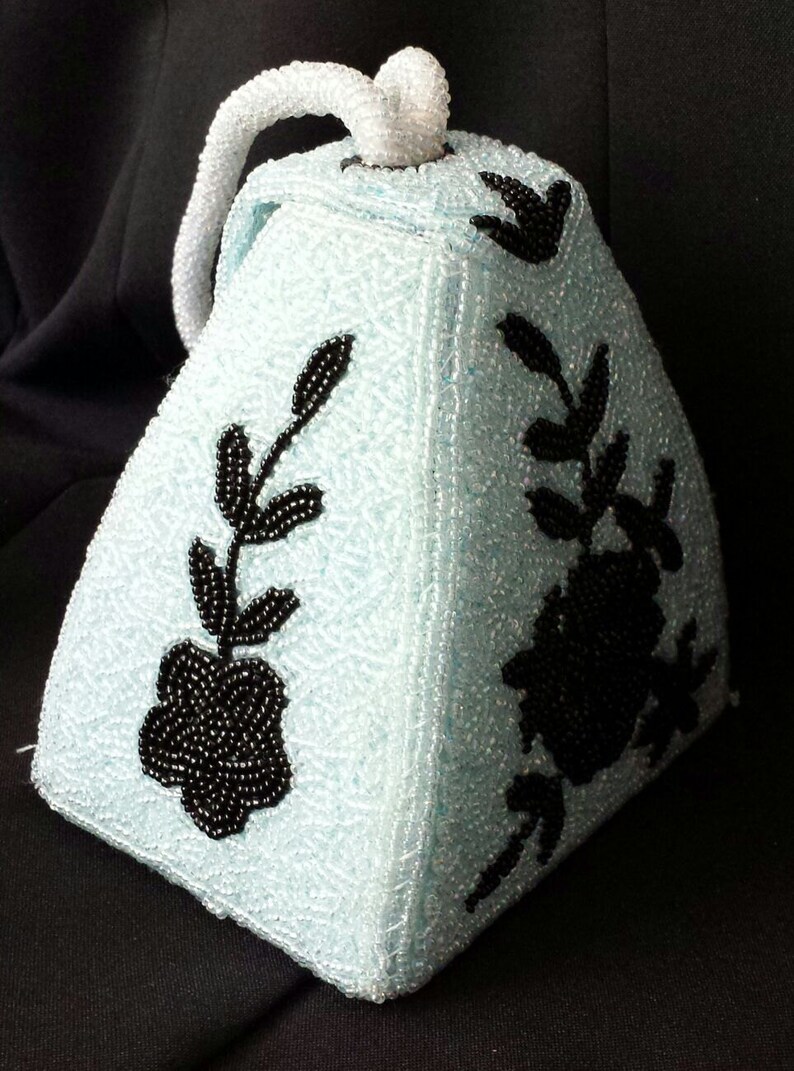 May include: A light blue beaded handbag with a black floral design. The bag has a handle and is shaped like a pyramid.