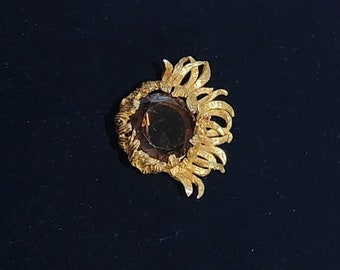 May include: A gold-toned brooch with a large, round, dark brown gemstone surrounded by a decorative, swirling design.