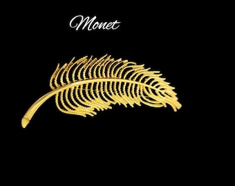 May include: A gold-colored feather-shaped brooch with the word "Monet" written in white script above it.