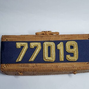 May include: A woven wicker clutch bag with a navy blue ribbon and gold numbers "77019" on the front.