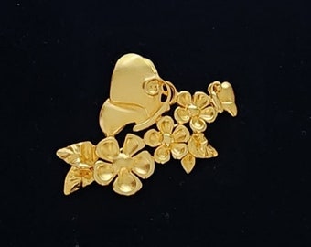 May include: Gold-toned brooch featuring a butterfly with floral accents. The butterfly is in a stylized design with wings that are shaped like hearts. The flowers are arranged around the butterfly and have a delicate, feminine look.