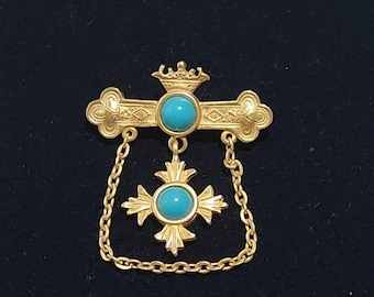 May include: A gold-toned brooch with a turquoise stone in the center. The brooch is in the shape of a cross with a crown on top and a chain hanging from the bottom.