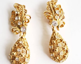 May include: A pair of gold tone dangle earrings with clear rhinestone accents. The earrings feature a teardrop shape with a filigree design and a smaller leaf-shaped design at the top.