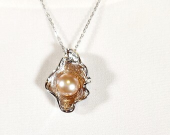 May include: A silver chain necklace with a pendant featuring a large, light pink pearl set in a silver and gold shell-shaped design.