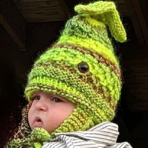 Pode incluir: Um chapéu de malha verde e amarelo com uma parte superior pontiaguda e abas para as orelhas. O chapéu é feito com um fio grosso e tem um padrão texturizado. O chapéu é usado por um bebê.
