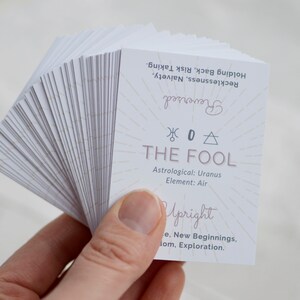 May include: A deck of tarot cards with a white background. The top card is The Fool, with the astrological symbol for Uranus and the element Air. The card is upright and shows the meaning of new beginnings, freedom, and exploration. The reversed meaning is holding back, risk taking, recklessness, and naivety.