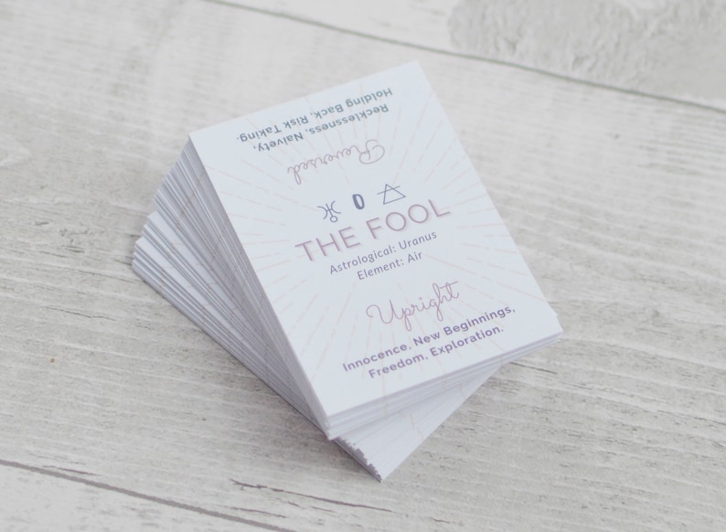 May include: A stack of white cards with a pink and gold design. The top card features the text "THE FOOL" with a symbol and the number "0". The card also includes the text "Astrological: Uranus, Element: Air, Upright, Innocence. New Beginnings. Freedom. Exploration."