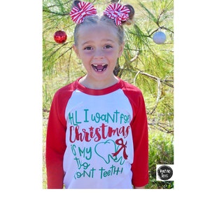 May include: A white and red long-sleeved shirt with a green and red graphic that says "All I want for Christmas is my two front teeth!" The shirt has a red and white bow on each side of the head.
