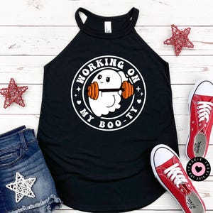May include: Black halter top with the text "WORKING ON MY BOO-TY" and a cartoon ghost lifting weights. The top is paired with denim shorts and red sneakers. Decorative red star ornaments are placed around the clothing.