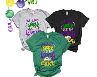 May include: Three t-shirts with the text "I'm just here for the king cake" in purple and green lettering. The shirts are in grey, black, and green.