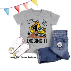 May include: A gray t-shirt with the text "I'm 4 & Digging It" and a cartoon excavator. The excavator is yellow with black details and is digging in brown dirt. The shirt is on a white background with a pair of white sneakers and blue jeans.