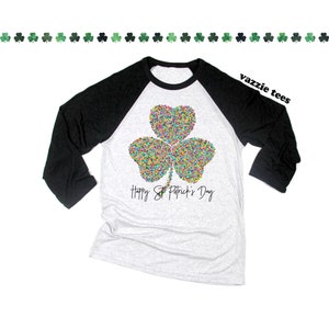 K&ouml;nnte beinhalten: Ein schwarz-wei&szlig;es Baseball-T-Shirt mit einem bunten Kleeblatt-Design und dem Text "Happy St. Patrick's Day".