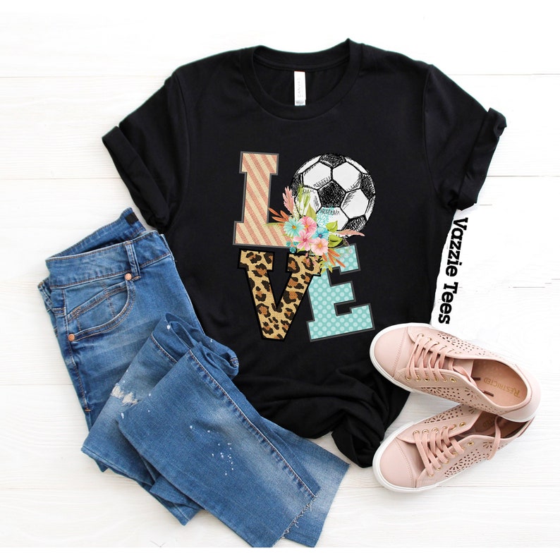 May include: Black t-shirt with a graphic of the word "LOVE" in colorful letters with a soccer ball and floral accents. The text "Vazzic Tees" is on the side of the shirt.
