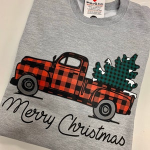 May include: A gray sweatshirt with a red and black plaid truck carrying a green and black plaid Christmas tree. The text "Merry Christmas" is written in black cursive below the truck.