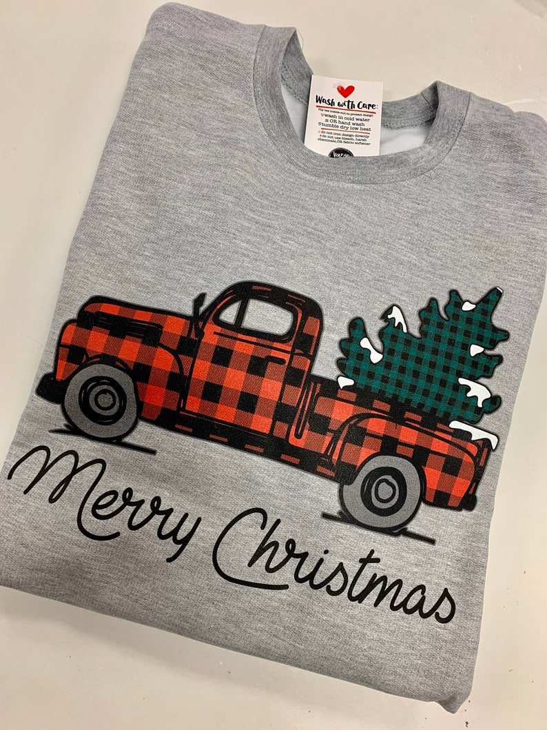 May include: A gray sweatshirt with a red and black plaid truck carrying a green and black plaid Christmas tree. The text "Merry Christmas" is written in black cursive below the truck.