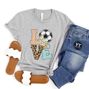 May include: Gray t-shirt with a graphic of the word "LOVE" with a soccer ball and floral accents. The letters are decorated with different patterns, including leopard print, stripes, and polka dots.