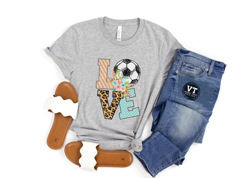 May include: Gray t-shirt with a graphic of the word "LOVE" with a soccer ball and floral accents. The letters are decorated with different patterns, including leopard print, stripes, and polka dots.