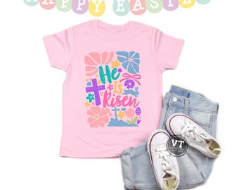 May include: Pink t-shirt with a colorful graphic design that says "He is risen" with a cross and flowers. The shirt is laid out on a white background with a pair of blue jeans and white sneakers.