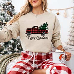 May include: A beige sweatshirt with a red and black plaid truck carrying a green Christmas tree. The text "Merry Christmas" is printed below the truck. The model is wearing red and black plaid pajama pants.