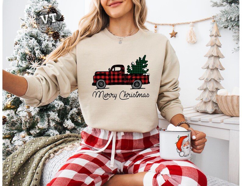 May include: A beige sweatshirt with a red and black plaid truck carrying a green Christmas tree. The text "Merry Christmas" is printed below the truck. The model is wearing red and black plaid pajama pants.