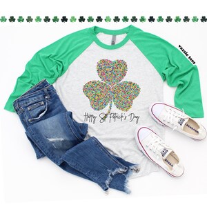 K&ouml;nnte beinhalten: Ein hellgraues und gr&uuml;nes Raglan-Shirt mit einem bunten Kleeblatt-Design und dem Text "Happy St. Patrick's Day".